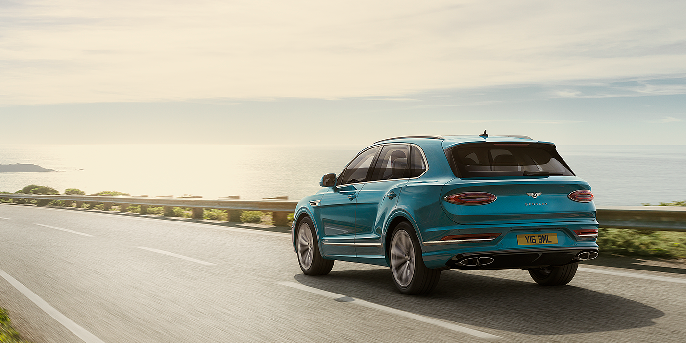 Jack Barclay Bentley Bentayga Azure luxury SUV rear three quarter in Topaz Blue by Mulliner paint, driving on an ocean road.