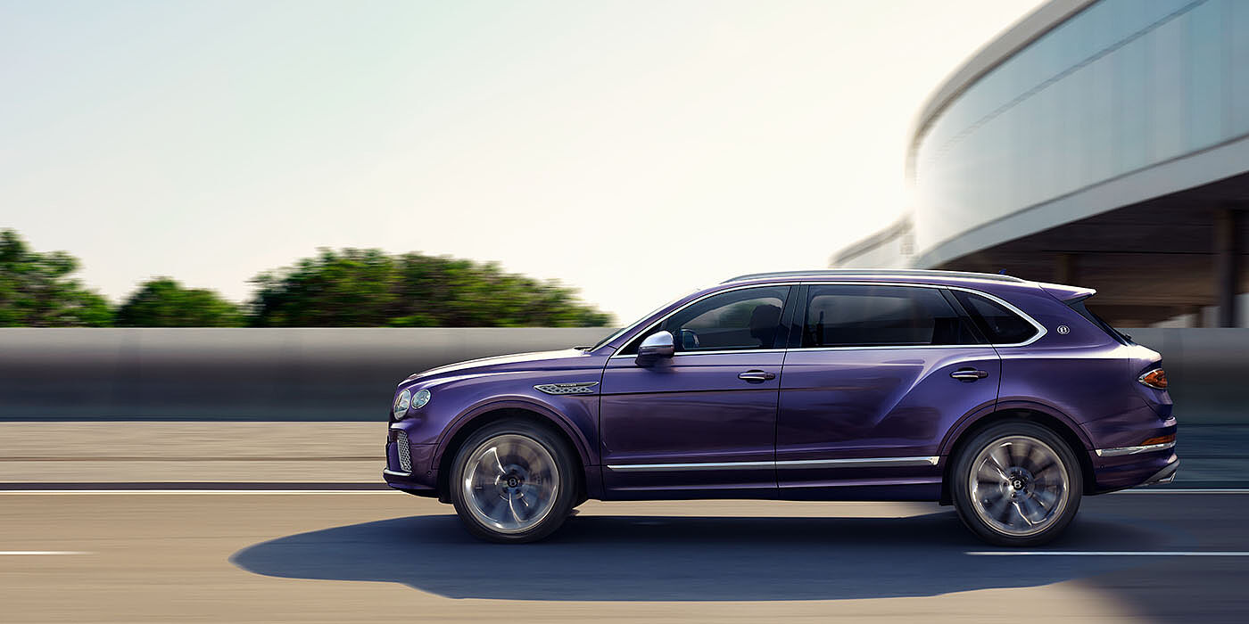 Jack Barclay Bentley Bentayga Extended Wheelbase Mulliner luxury SUV profile in Tanzanite Purple paint, driving on an country road past trees.