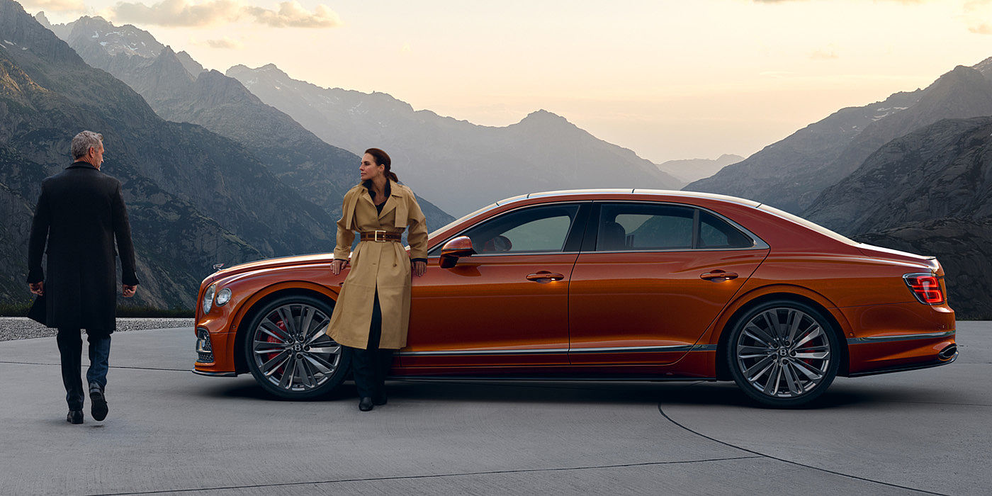 Jack Barclay Bentley Flying Spur Speed parked in Orange Flame coloured exterior parked, with mountainous background and two people in view.