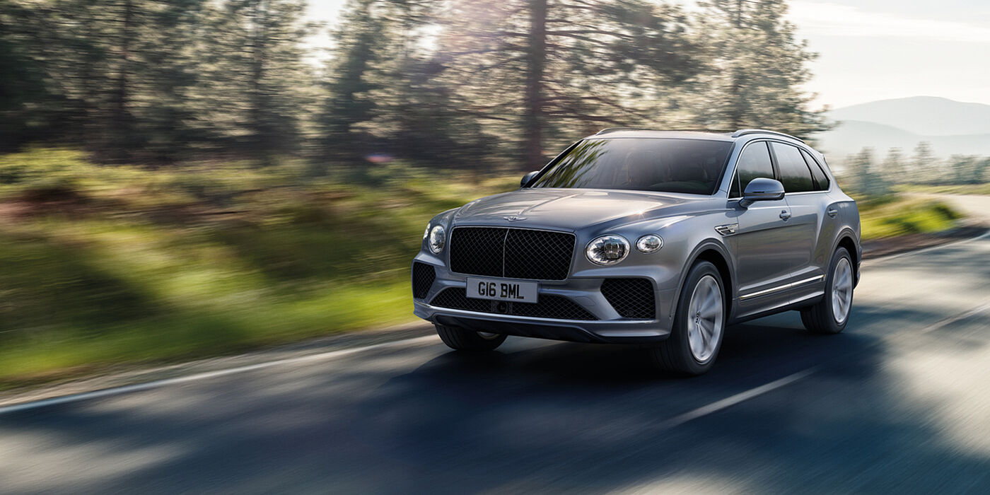Jack Barclay Bentley Bentayga luxury SUV front three quarter in Moonbeam paint, driving on an country road past trees.