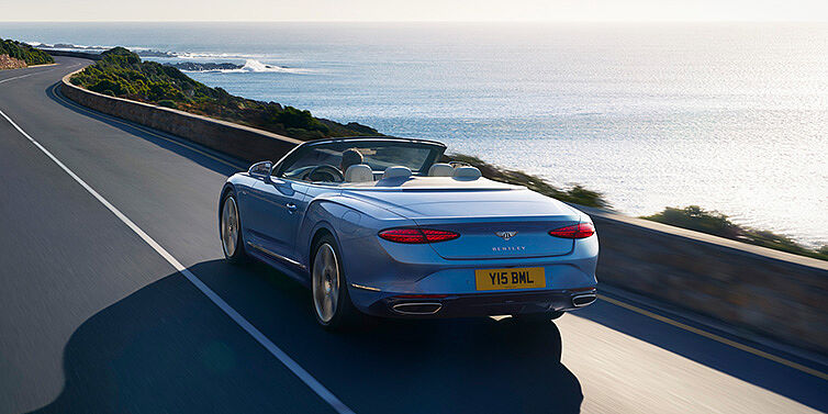 Jack Barclay New Bentley Continental GTC Azure convertible rear view in Light Windsor blue paint with 22 inch ten spoke wheels driving dynamically on an ocean road in the sunshine