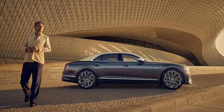 Jack Barclay Bentley Flying Spur Mulliner sedan in Rose Gold over Magnetic duotone paint parked by a curved wall with person in suit jacket standing on the left